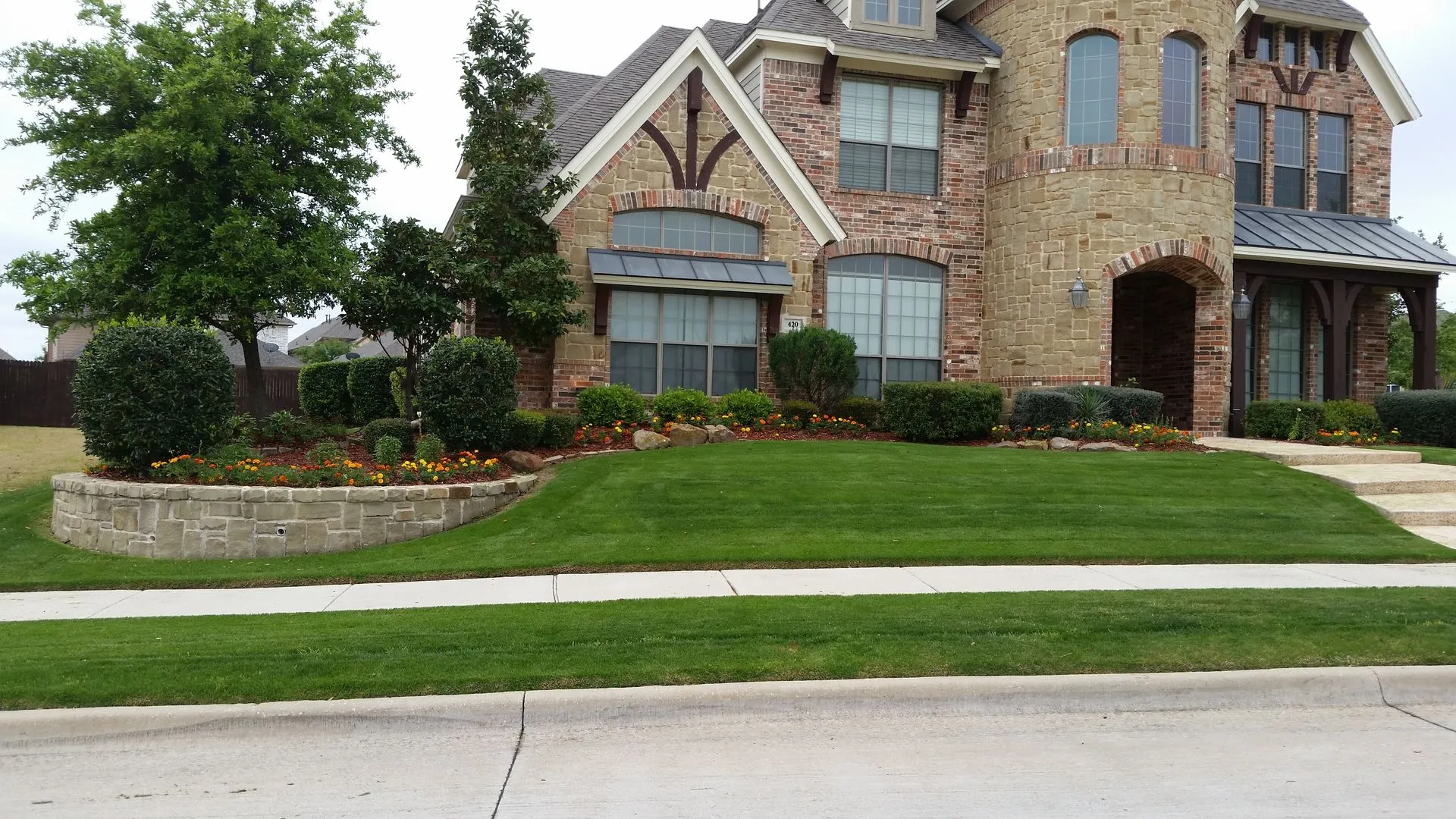 Curved stone retaining wall bed with shrubs and marigolds in front of a stone home