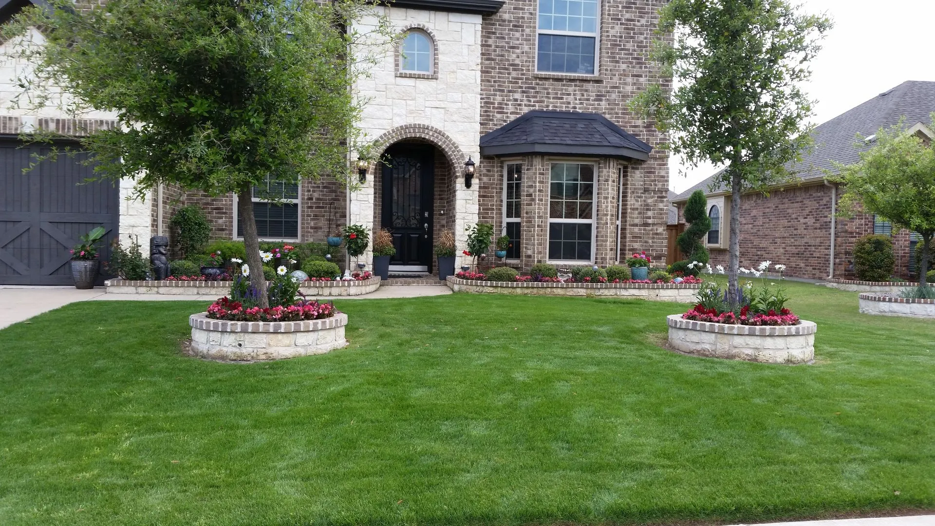 Stone-bordered tree rings and front beds full of flowers across a green lawn